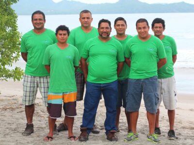 Meet the Team A photo of the entire maintenance team of Special Places of Costa Rica. Two rows of smiling men, all wearing lime-green 'Special Places' t-shirts and posing on a beautiful tropical beach.